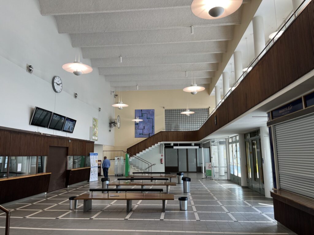 A spacious indoor area with high ceilings, large wall clocks, seating benches, a staircase to the upper level, and a few people walking. There are several display screens and a closed concession stand.