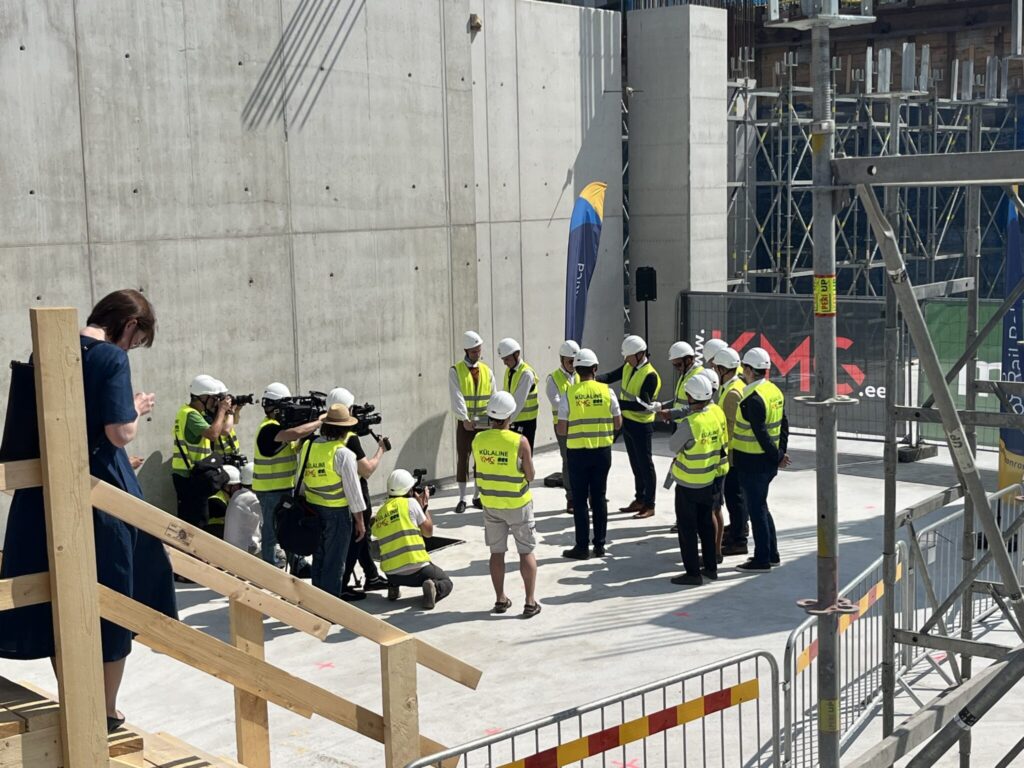 A group of construction workers in yellow safety vests and white helmets gather on a concrete site with a few people taking photos. Wooden stairs and construction scaffolding are visible at the edges of the photo