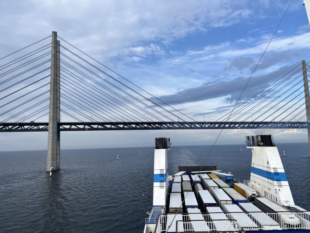 A ferry transporting trucks sails under a suspension bridge stretching across a body of water on a partly cloudy day.