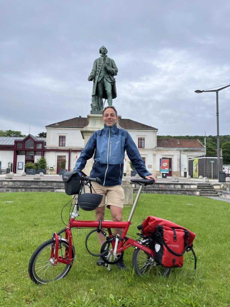 Jon Worth, a middle aged man, standing with a red bicycle in front of a statue on a cloudy day, with a building (Montbard Station) and green lawn in the background.