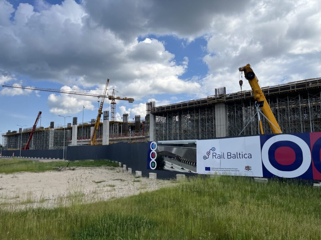 A construction site for the Rail Baltica project is shown, with cranes and scaffolding in action under a partly cloudy sky. A blue and white sign on a fence reads 