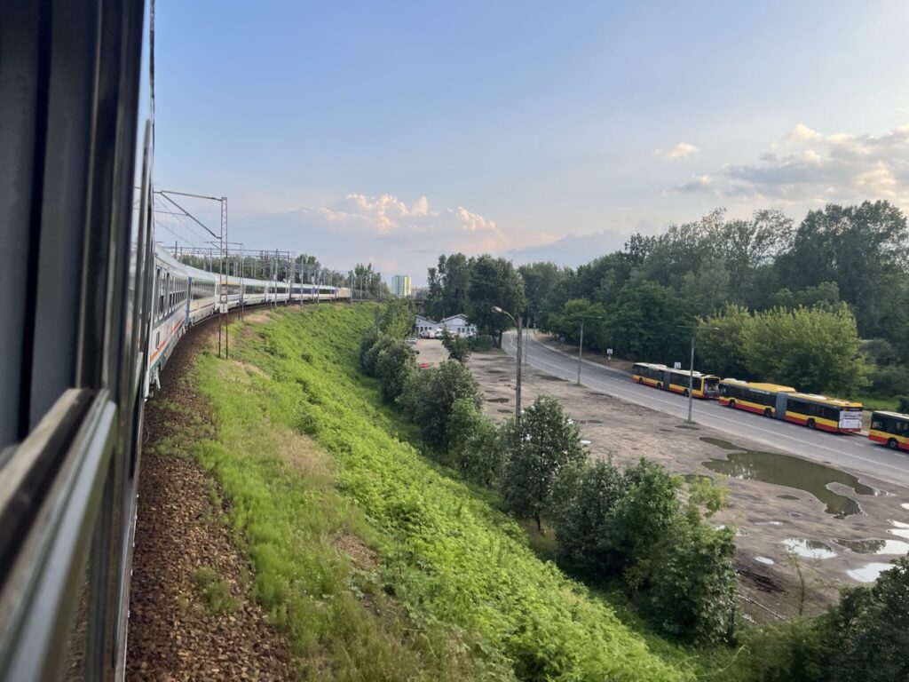 View from a train window showing the locomotive and first carriages of the train curving along an embankment covered with greenery. Below, a road with several yellow buses is surrounded by trees and patches of wet ground reflecting the sky. In the distance, the horizon features more greenery and a partly cloudy sky.