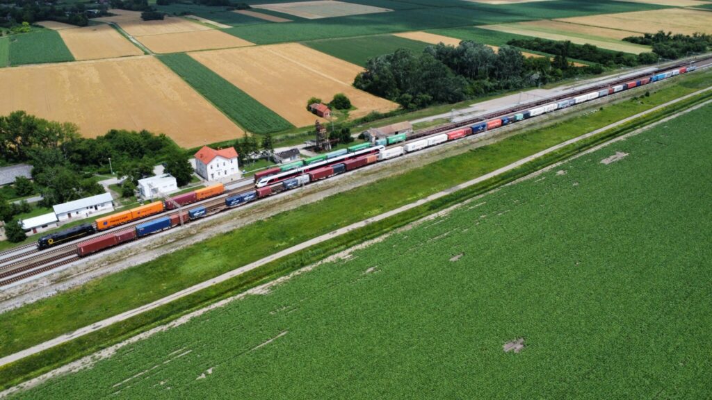 Aerial view of a long freight train with colorful containers. It is stopped at a station surrounded by grassy fields and farmland. The train tracks run alongside a narrow road. Several buildings, including a white station building with a red roof, are positioned near the tracks amidst the countryside landscape.