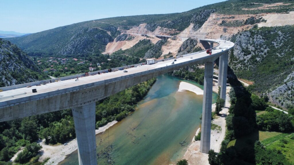 Aerial view of a high, elevated concrete bridge under construction spanning a lush valley with a river below. The bridge winds through mountainous terrain with construction areas visible on nearby hillsides. Sparse constuction traffic, including a truck, can be seen on the bridge. Dense greenery surrounds the riverbanks.