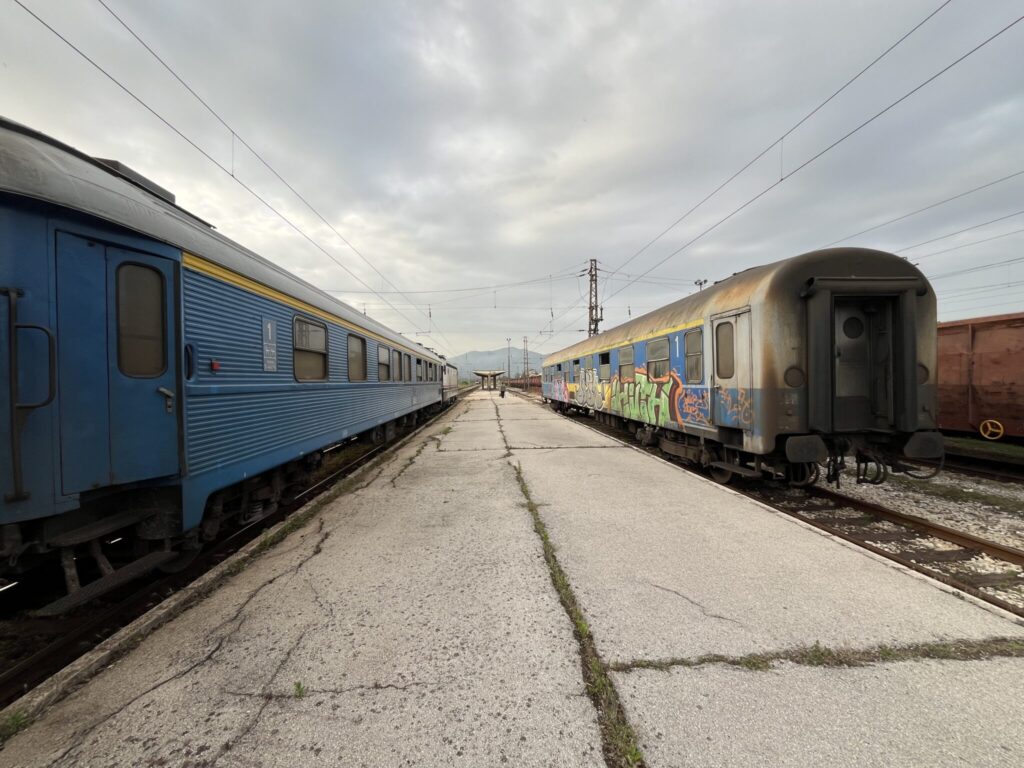 Two old train cars are on parallel tracks on a cloudy day. A locomotive is just visible hauling the left one. The left train car is blue, while the right train car has blue and white graffiti on its side. The ground between the tracks is cracked and worn. Power lines are visible overhead with poles lining the tracks in the background.