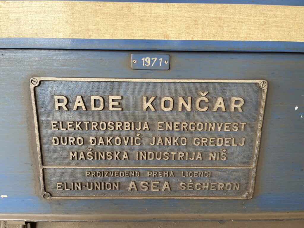 A metallic plaque on a blue background. The plaque has raised text in Serbian: 