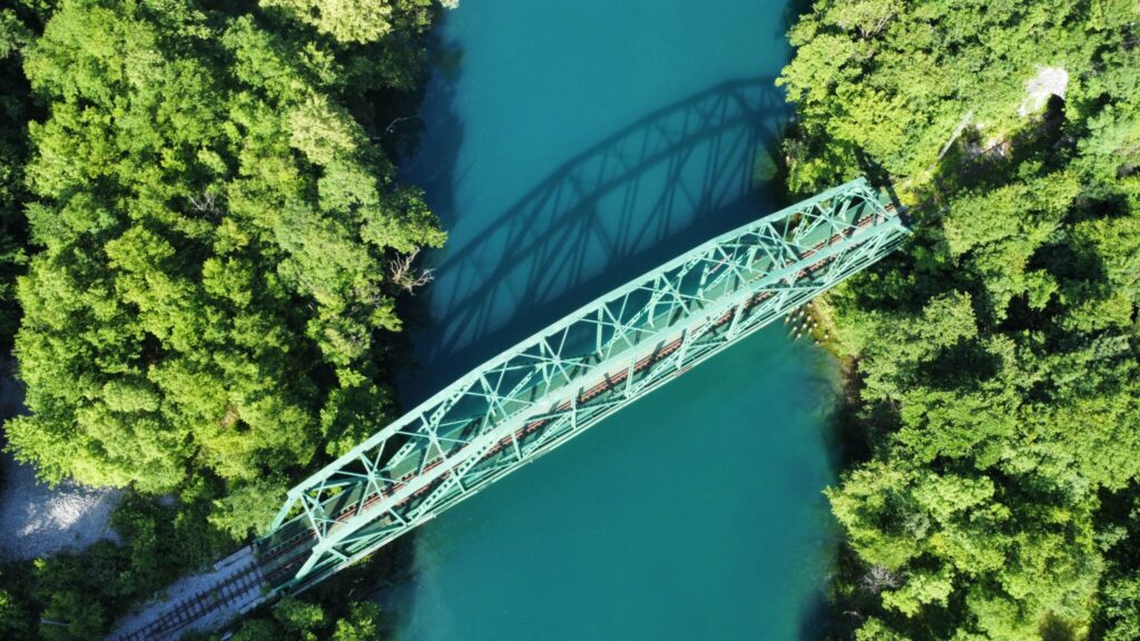 Aerial view of a green steel truss bridge spanning over a blue-green river surrounded by dense green foliage. The bridge casts a clear shadow on the water below. The natural scenery is vibrant with lush greenery and clear waters. There are no vehicles or people visible on the bridge.