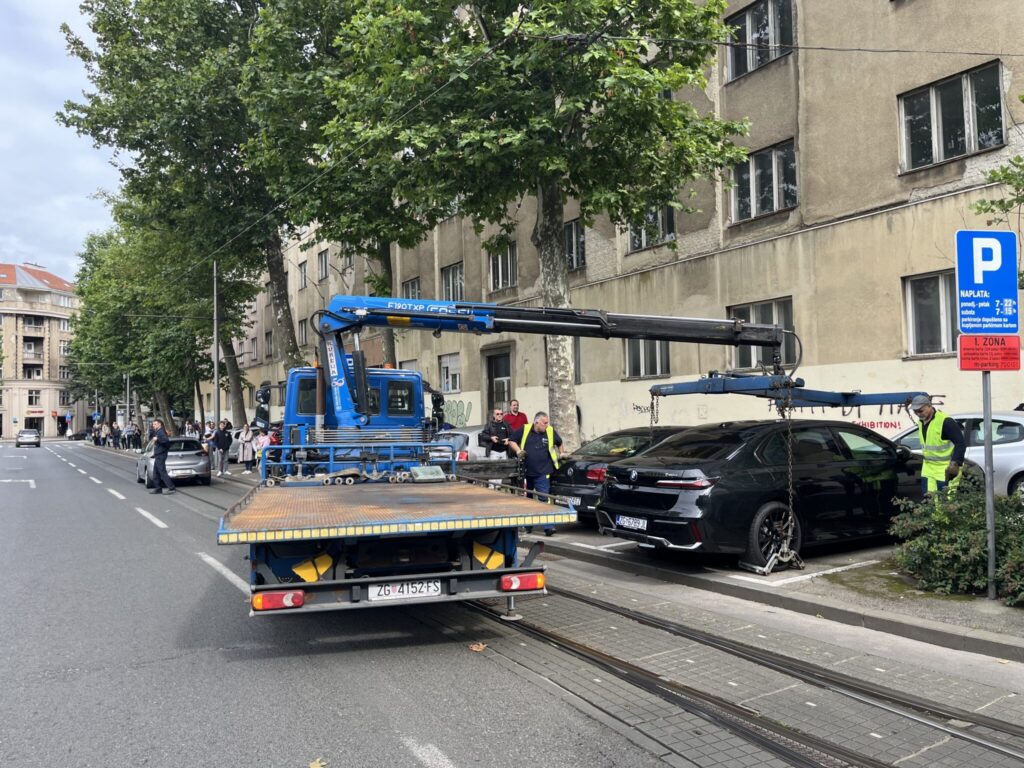 A tow truck with a blue crane arm is lifting a black car from the sidewalk onto its flatbed. A few workers in neon vests assist. The scene takes place on a city street lined with parked cars and trees in front of an aged beige building. A blue parking sign with white text is visible.