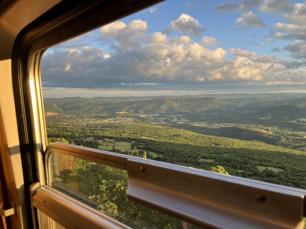 View from a train window of a vast landscape with green hills and valleys under a sky with scattered clouds, illuminated by warm sunlight. The scene captures the rolling terrain, with some distant sparse structures and villages, reflecting a serene countryside journey.