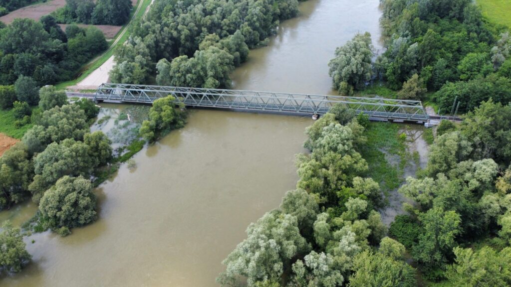 An aerial view of a metal truss bridge spanning a wide, muddy river surrounded by dense, green trees. The river is high and surrounds some trees. The bridge is used for rail transport.