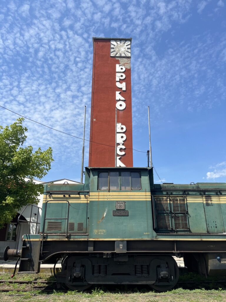 A vintage green and yellow diesel locomotive is stationed on railway tracks. Behind it stands a tall, rectangular, red brick tower with white Cyrillic letters reading Brčko and a clock near the top. A tree with green leaves is on the left side, and the sky is mainly clear with scattered clouds.