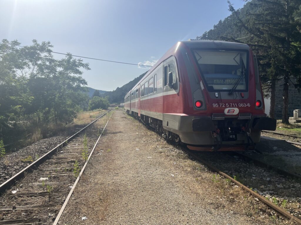 A red and grey train is stationary on a track surrounded by greenery and mountains under a clear blue sky. The train has the number 