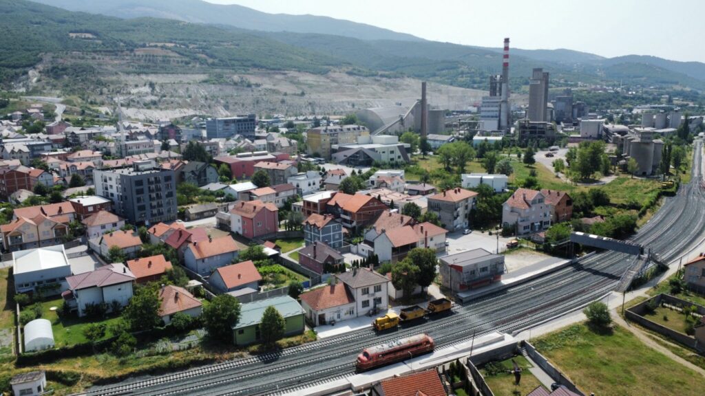 Aerial view of a residential area with rows of houses featuring red-tiled roofs. Two trains are stationary on the train tracks parallel to and in front of the houses. The smaller 2 axel train is yellow, the larger 6 axel one is dark red. An industrial complex with smokestacks is visible in the background, set against a backdrop of green hills and clear skies.