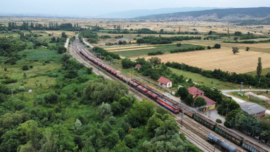 Aerial view of a train in the middle through a green agricultural landscape. The train is hauled by a red electric locomotive, and comprises numerous freight cars, and some assorted freight cars from other trains are visible in front of it. A small station building and a level crossing are visible near the tracks. Rolling hills and a distant town are seen under a cloudy sky.