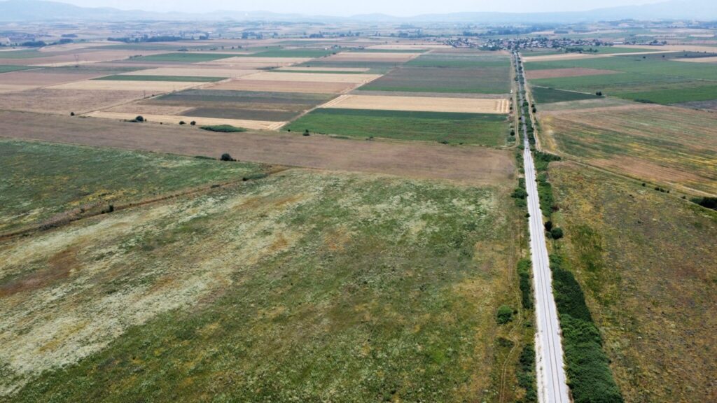 An aerial view of a vast rural landscape with large, segmented agricultural fields of various shades of green and brown. A single track railway line cuts through the fields, leading towards a distant town. Hazy mountains can be seen in the background under a light blue sky.