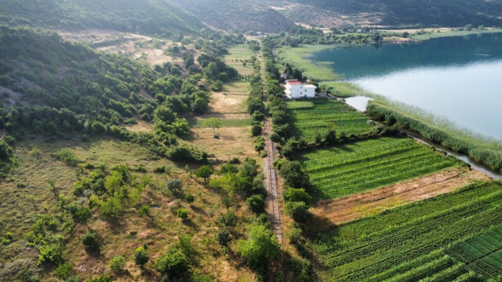 Aerial view of a lush, green landscape with a lake on the right. A white house with a red roof sits near the lake, surrounded by dense vegetation and farmlands. An abandoned railway line, single track, runs through the centre, flanked by trees and fields. Rolling hills in the background are covered in greenery.