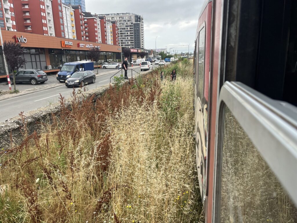 View from a train running through an urban area with tall grasses and weeds along the tracks. On the left, a busy street runs parallel, with several vehicles passing by. Modern apartment buildings and a shop or mall with a visible Silvio sign are in the background. It's a cloudy day.