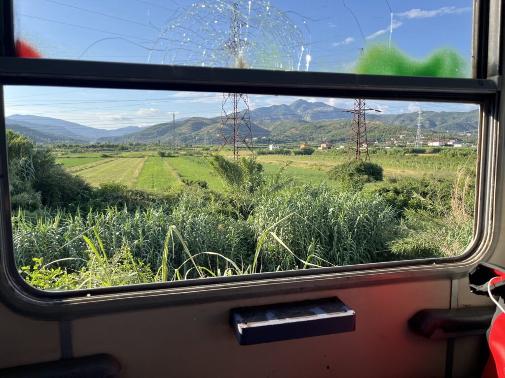 View through a train window with a cracked upper pane. The landscape outside features lush green fields, dense vegetation, and distant mountains under a clear blue sky. Red and green marks are visible on the window. The train interior includes a small table handle and a corner of a seat.