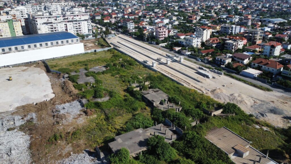 Aerial view of a railway construction site with large concrete slabs for future platforms and equipment on site, surrounded by urban buildings. To the left are unfinished structures and to the right, dense residential buildings with red roofs. A clear demarcation of vegetation and bare land is visible to the left centre of the picture