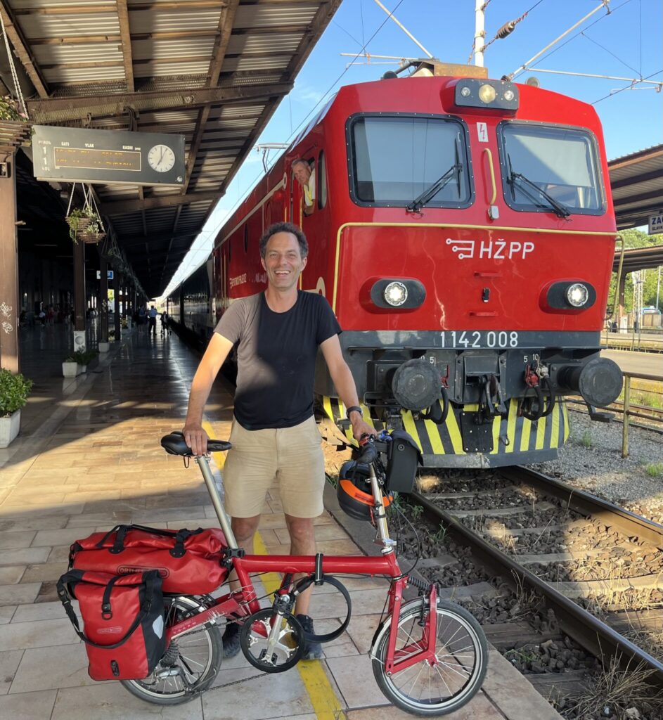 Jon Worth, a middle aged man, stands on a train platform smiling, holding a red foldable bicycle. Behind him is a red train with 