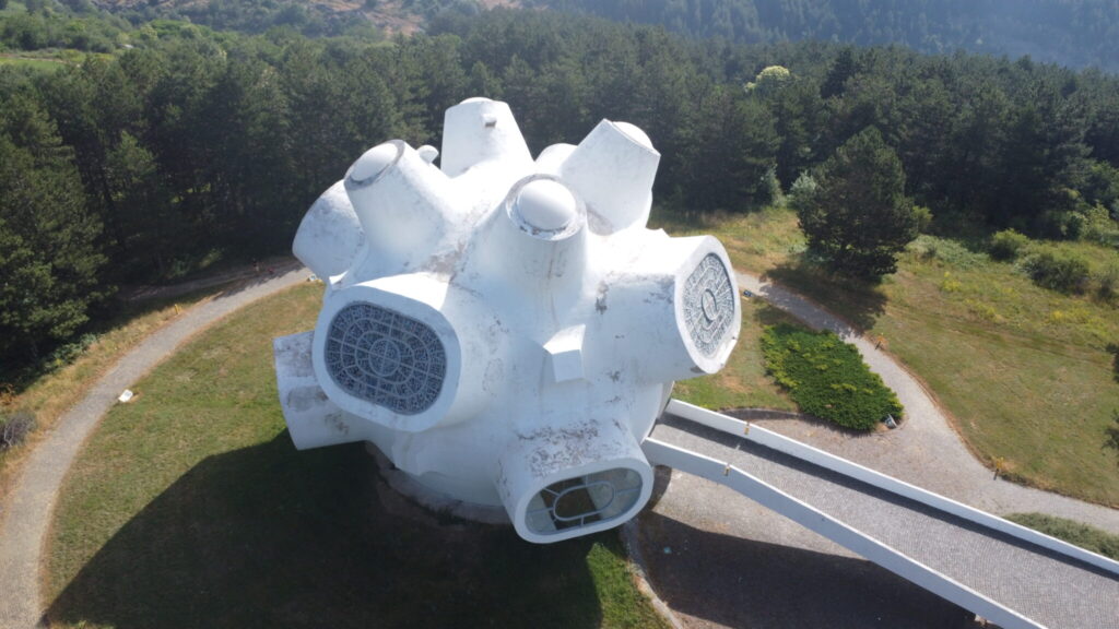 Aerial view of the Ilinden Monument in Kruševo, Macedonia, a large, white, abstract molecular shape structure with circular windows, surrounded by trees and pathways.