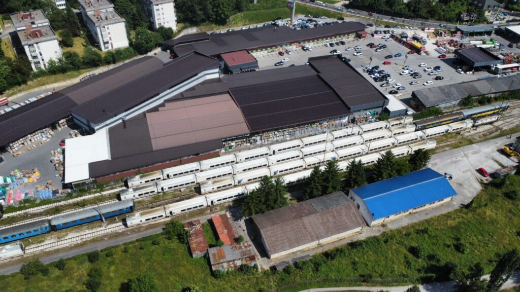 Aerial view of a large building complex next to multiple railway tracks, with several parked white Talgo trains and a parking lot filled with cars. Surrounding area includes greenery and additional buildings.