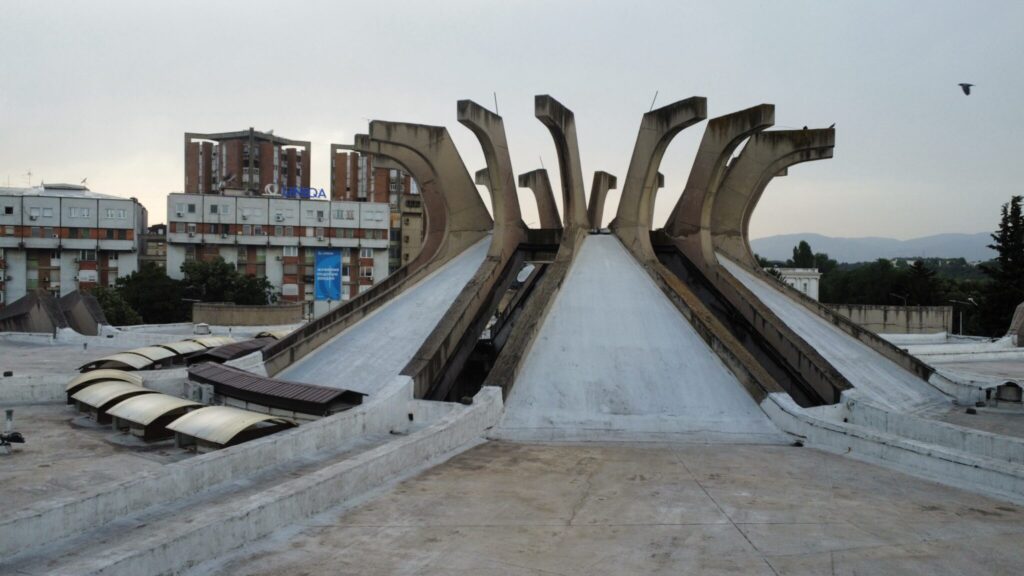 Central Post Office Skopje. A large, unique, concrete building structure with multiple pointed, curved supports on its roof. Surrounding concrete buildings and overcast sky are also visible.