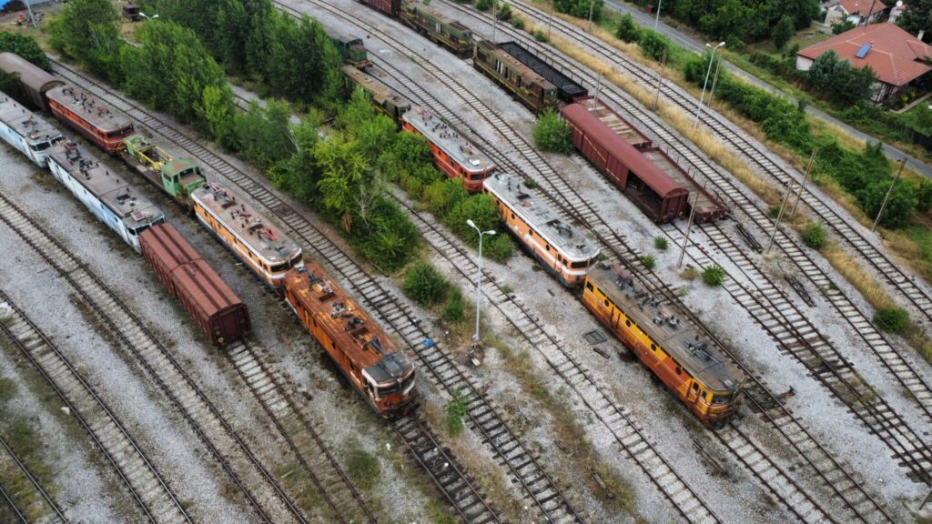 Aerial view of a train yard with rusted and overgrown railway tracks featuring multiple old electric and diesel locomotives. The diesels are mostly green, the electrics either orange or blue. Trees and vegetation grow through the tracks.